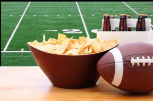 Chips, football and Six Pack of Beer on a table in front of a big screen TV with a Football field. Great for Super Bowl themed projects. Horizontal format.
